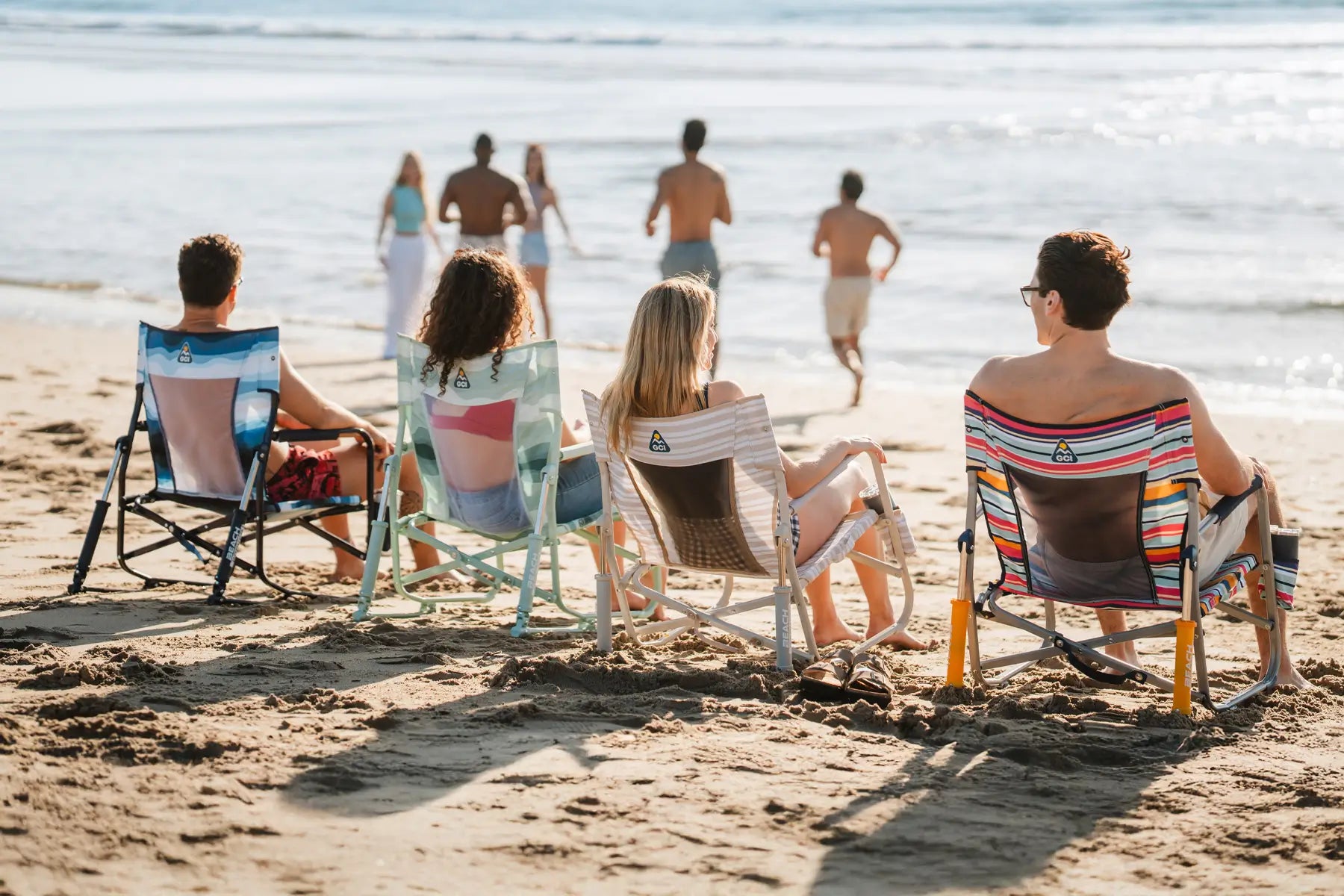 Four people sitting in Beach Rockers facing the ocean, with others walking along the shoreline.