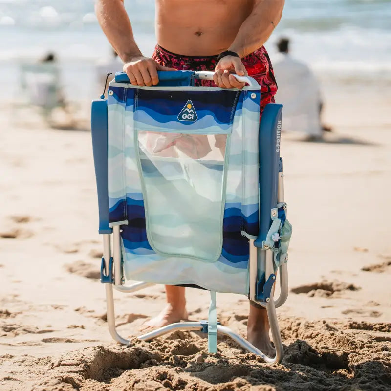 Man holding a folded Bi-Fold Beach Chair with blue wave patterns and a mesh back on the sand.