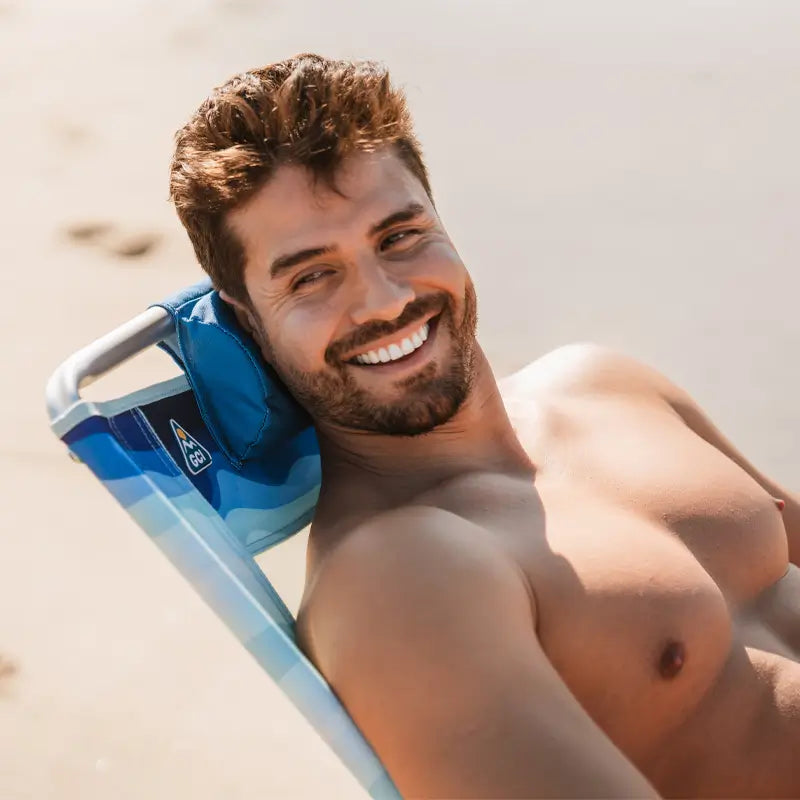 Close-up of a smiling shirtless man reclining in a Bi-Fold Beach Chair with blue wave patterns and a blue headrest.