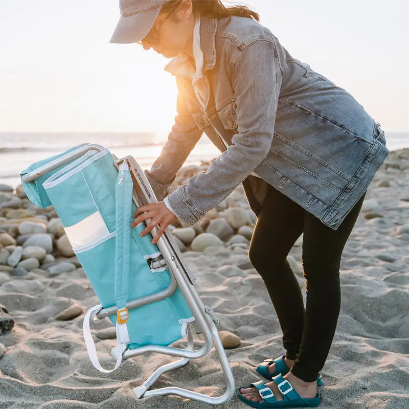 Woman wearing a denim jacket and blue sandals setting up a turquoise Bi-Fold Beach Chair on a rocky beach at sunset.