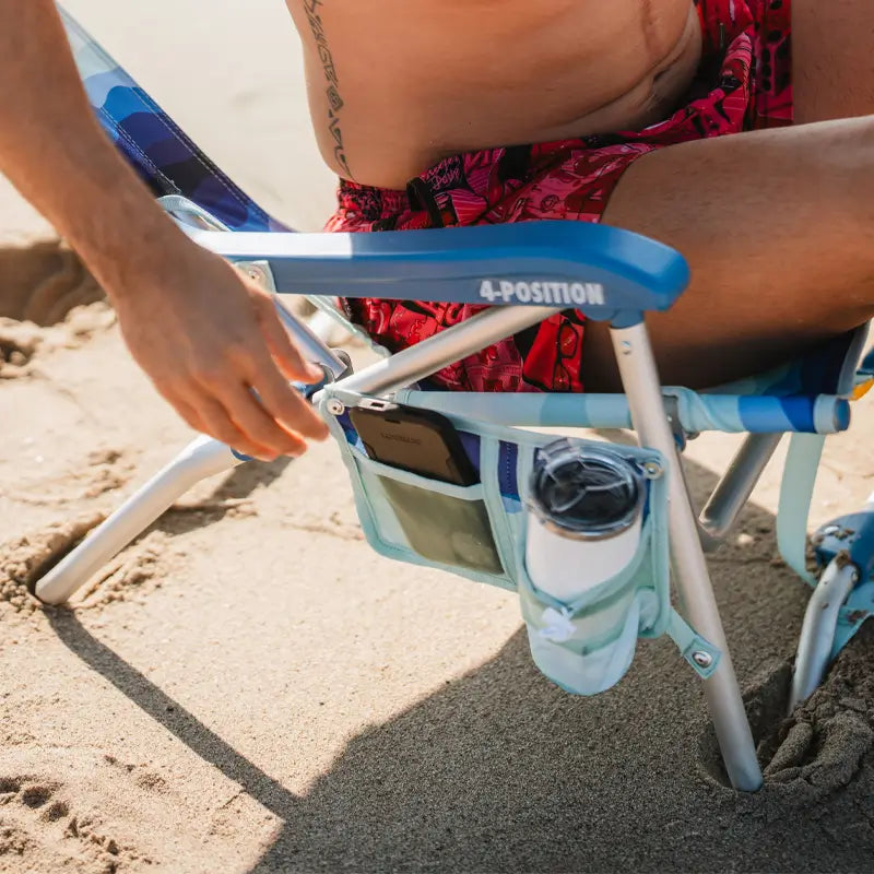 Man sitting in a Bi-Fold Beach Chair with a blue frame, storing a phone in the side pocket and a drink in the cup holder.