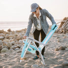 Woman wearing a denim jacket and hat, setting up a Bi-Fold Beach Chair on a sandy beach with rocks at sunset.