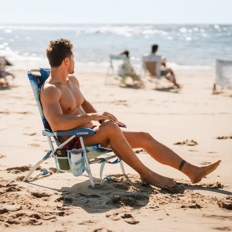 Man relaxing shirtless in a blue Bi-Fold Beach Chair on sandy shore, with drink and storage pouches attached to the armrest.
