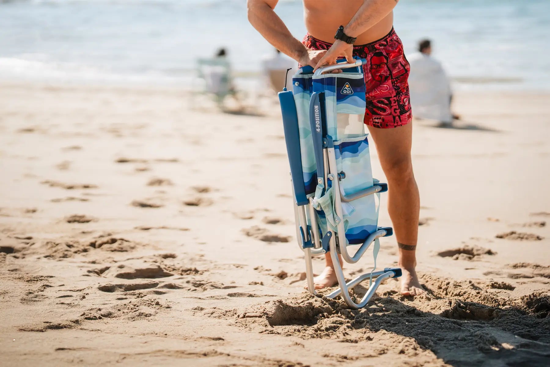 Man holding a folded Bi-Fold Beach Chair on a sandy beach with waves in the background.