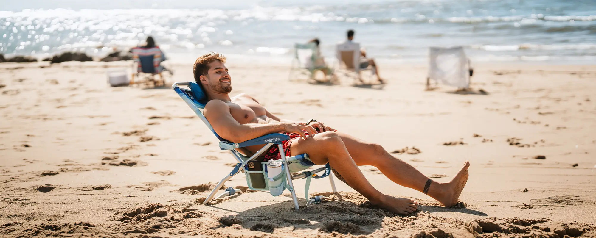 A shirtless man reclined on the Bi-Fold Beach Chair while relaxing on the beach. 