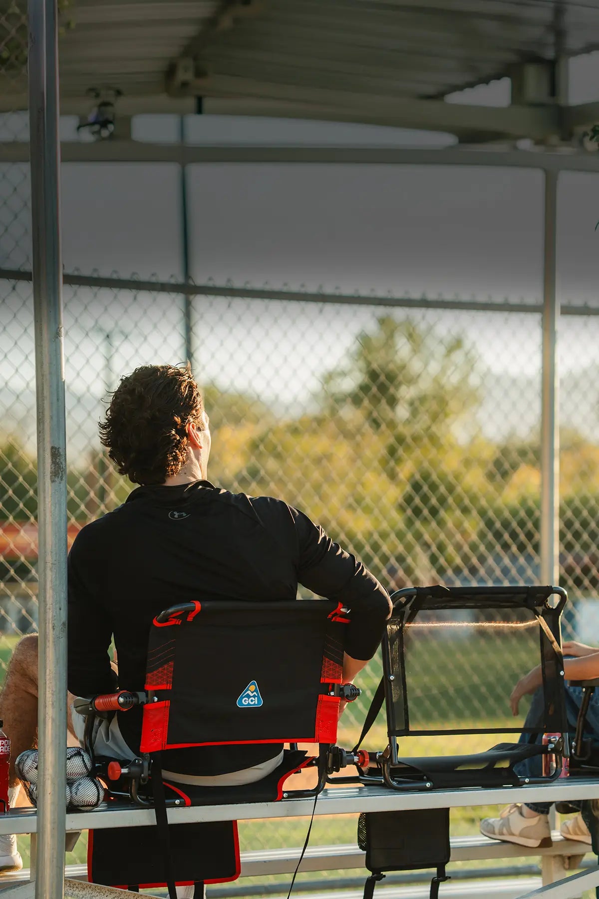 A person sitting on a black and red Big Comfort Stadium Chair, viewed from behind as they look toward a chain-link fence. The chair has a blue triangular logo on the backrest.