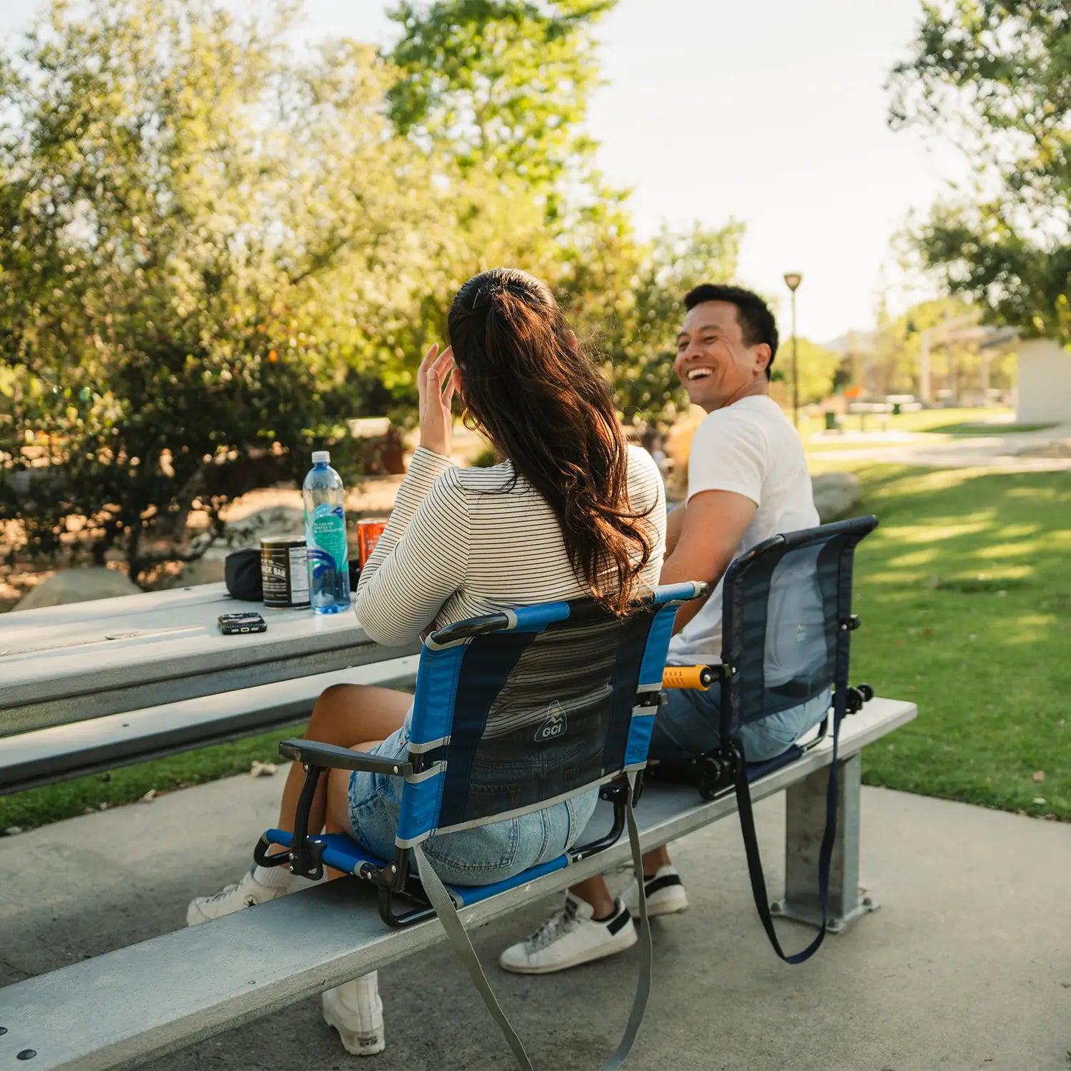 A couple having drinks together as they sit on a metal bench with their Big Comfort Stadium Chair and Stadium Rock-cliner. all-groups