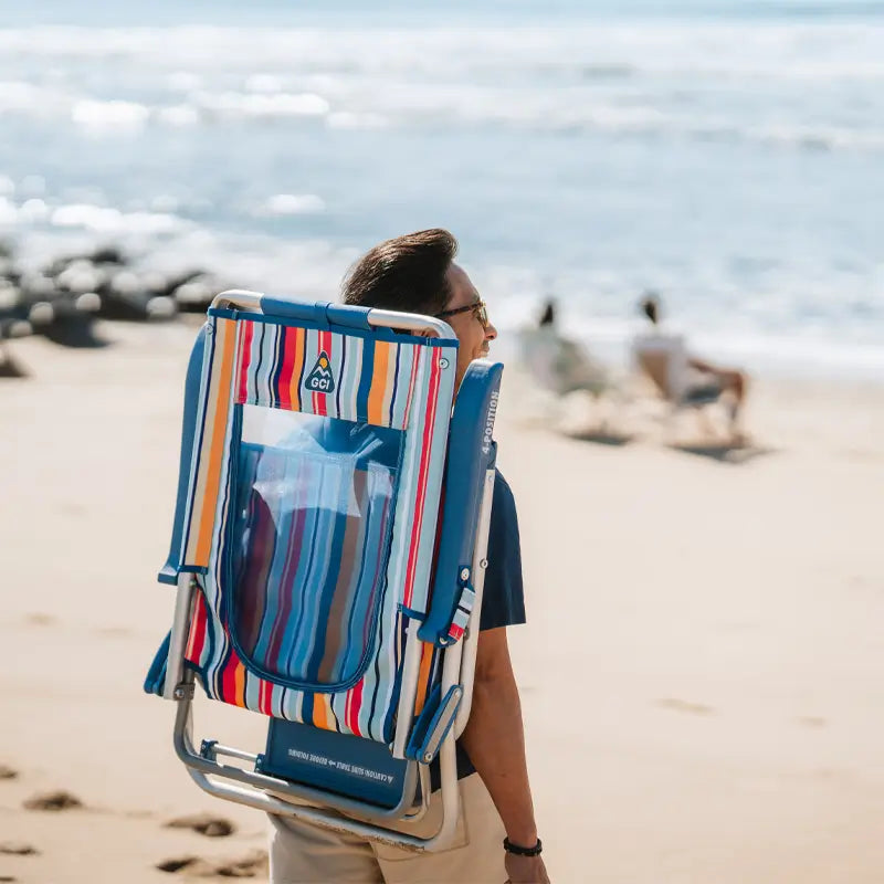 Man carrying a folded Big Surf beach chair with colorful stripes on his back while walking along the sandy beach.