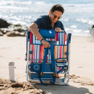 Man smiling while adjusting the straps on the back of a folded Big Surf beach chair with a colorful striped design.