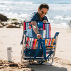 Man securing the backpack straps of a folded Big Surf beach chair with a blue headrest and striped pattern.