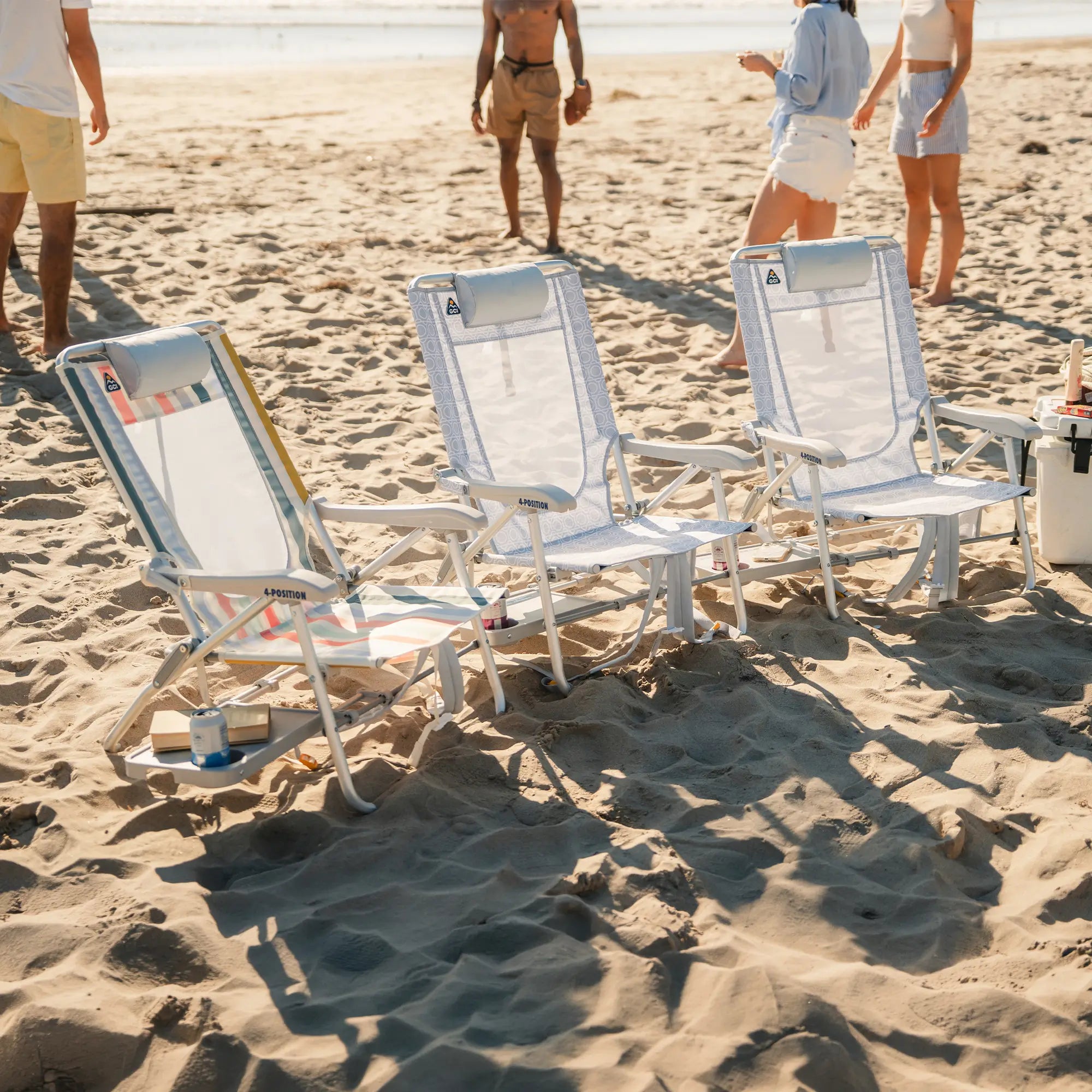 Three white beach lounge chairs with striped blue and yellow accents are set on a sandy beach, with people walking in the background near the water.