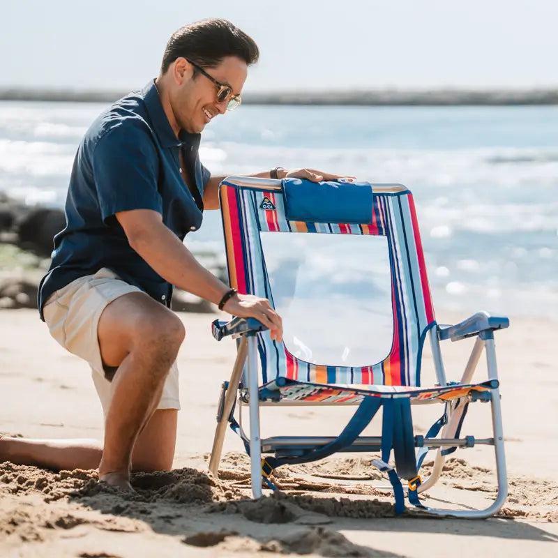 Man kneeling on sand, setting up a colorful striped Big Surf beach chair with a mesh back and blue headrest by the ocean.