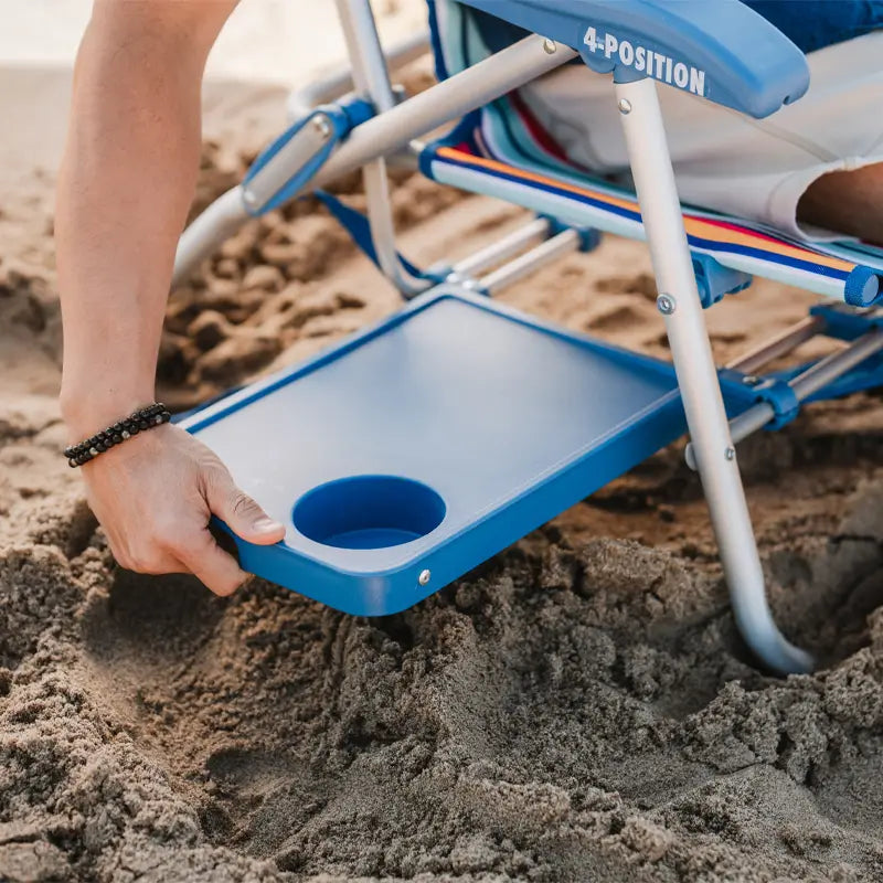 Close-up of hand pulling out the blue slide table with a cup holder from a Big Surf beach chair on sandy beach.