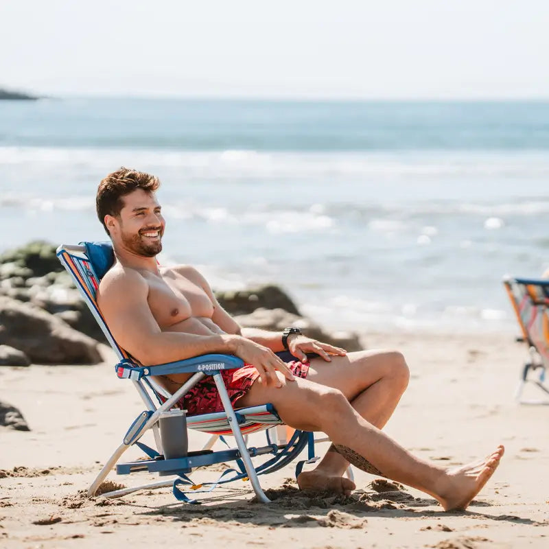 Shirtless man in red swim trunks reclining in a Big Surf chair with a cup in the slide table, smiling by the ocean.