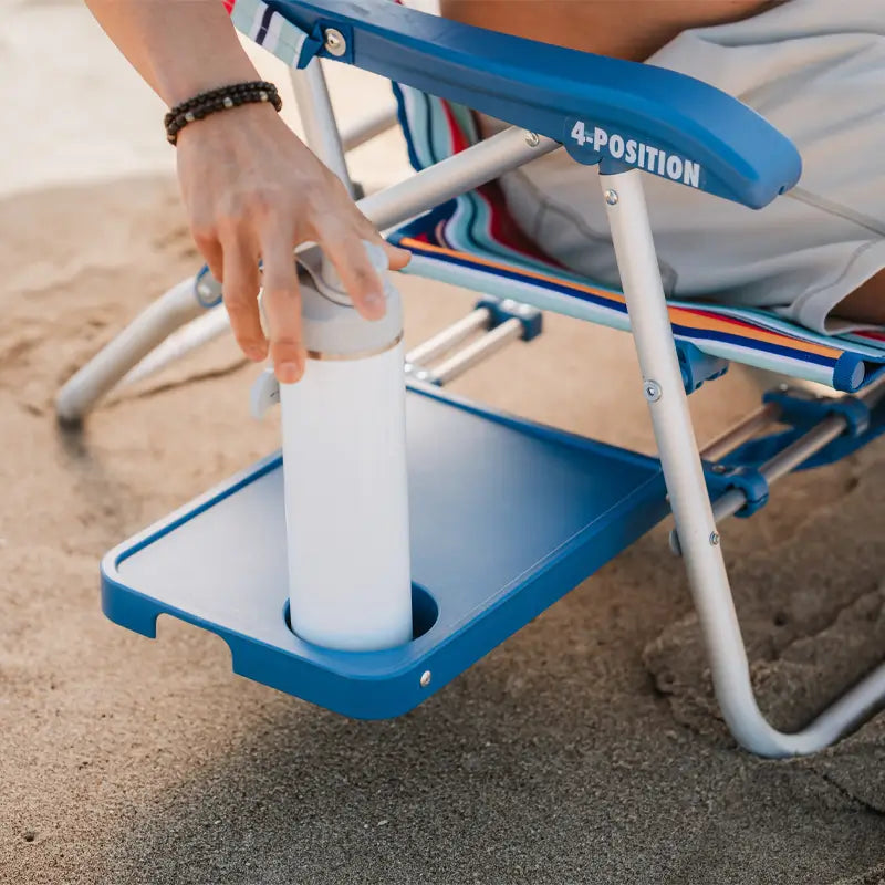 Hand placing a white bottle into the built-in slide table cup holder of a multi stripe Big Surf beach chair.