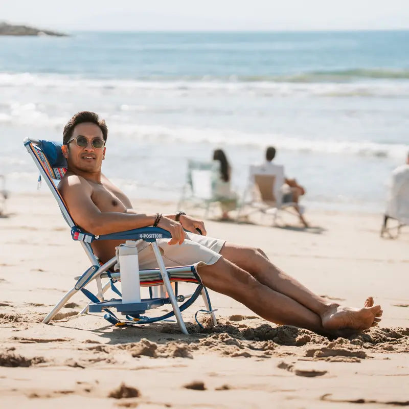 Man reclining in a multi stripe Big Surf chair with a slide-out table, relaxing on the beach facing the ocean.