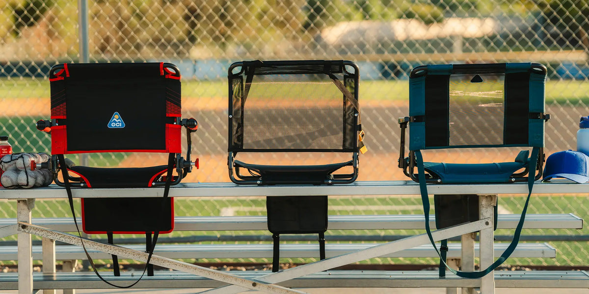 Three portable folding GCI stadium chairs in red, black, and blue are arranged on metal bleachers in front of a chain-link fence at a baseball field.