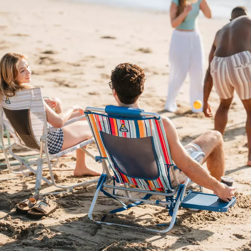 Man sitting in a Big Surf chair on the beach, holding a drink while chatting with a woman nearby.