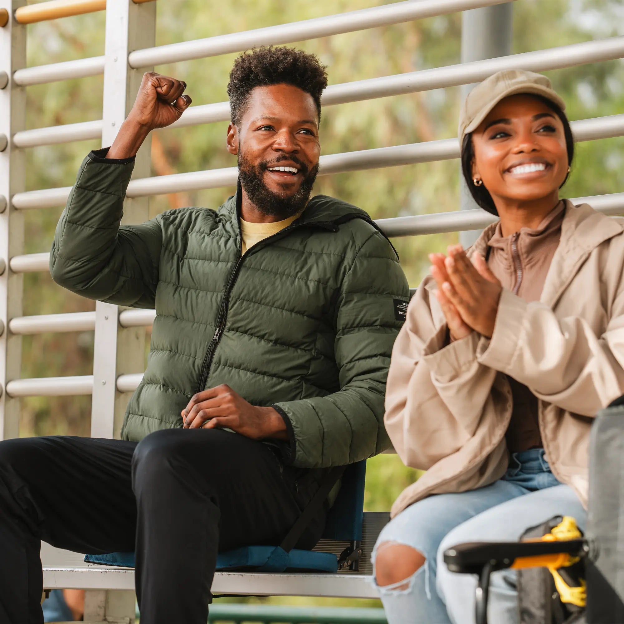 A man and woman cheer from metal bleachers while sitting on padded Bleacher Back seats, showing their comfort and support.