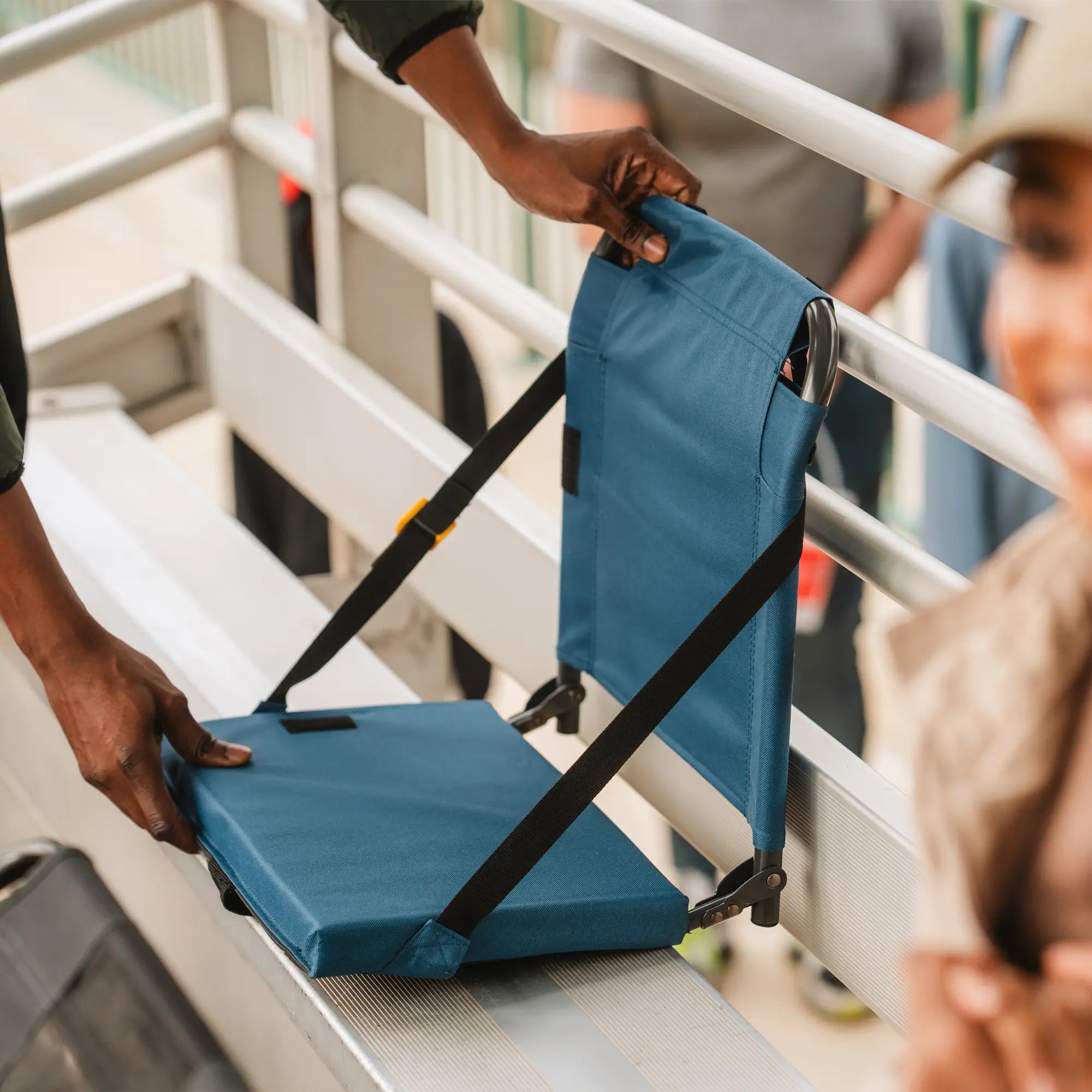 A person unfolds a blue Bleacher Back seat on metal bleachers, demonstrating its compact and portable design.