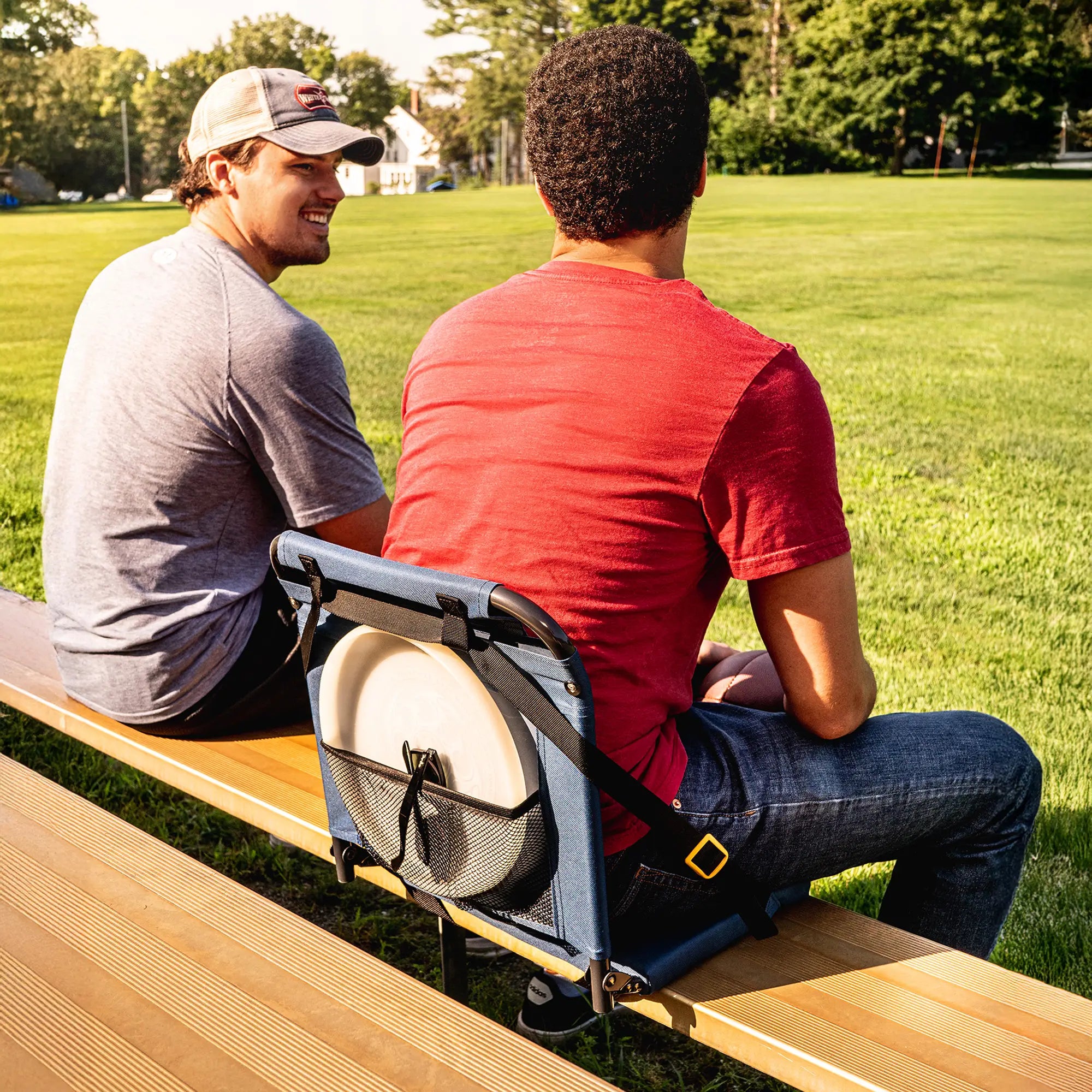 Two men sit on wooden bleachers with Bleacher Back seats, one storing a frisbee in the seat's back pocket, showcasing its storage feature.