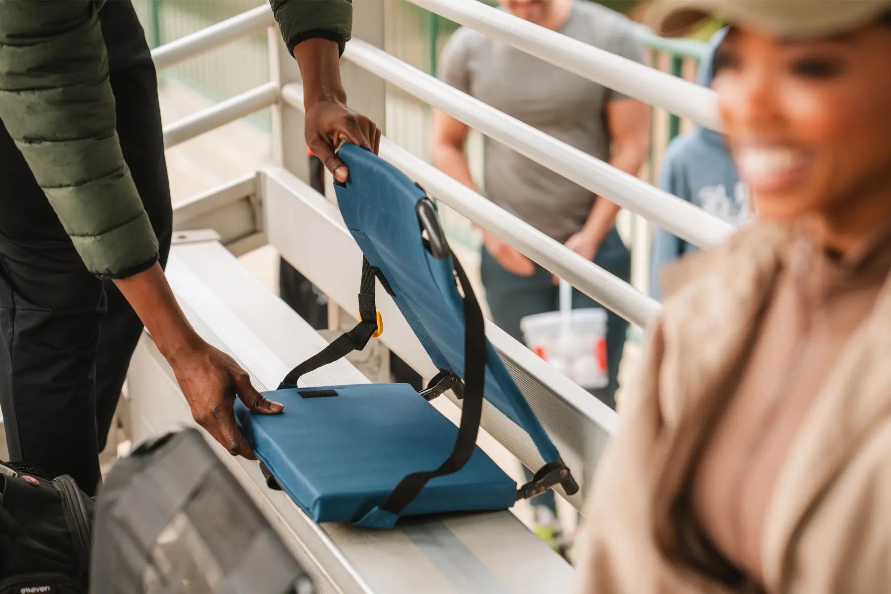 A person sets up a Bleacher Back seat on bleachers, with people socializing in the background, highlighting its convenience and comfort.