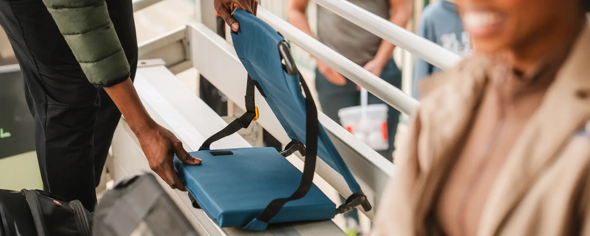 A person sets up a Bleacher Back seat on bleachers, with people socializing in the background, highlighting its convenience and comfort.