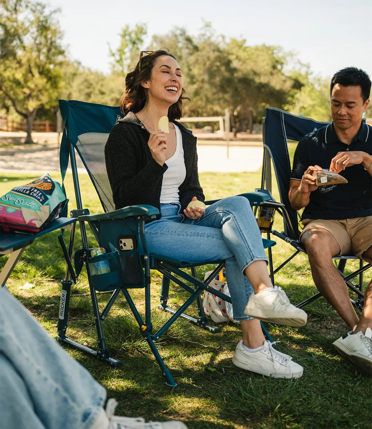 A woman sitting in a BreezeRocker at a park while eating a chip. 