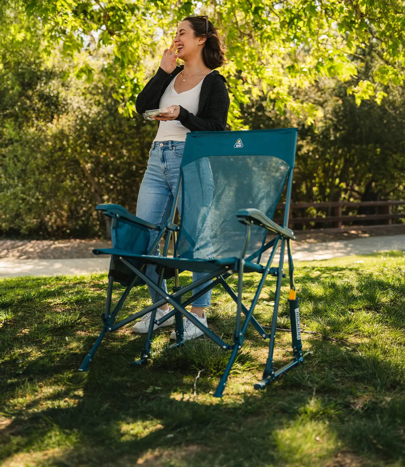 A woman standing eating a snack at a park while a teal BreezeRocker sits in front of her. 
