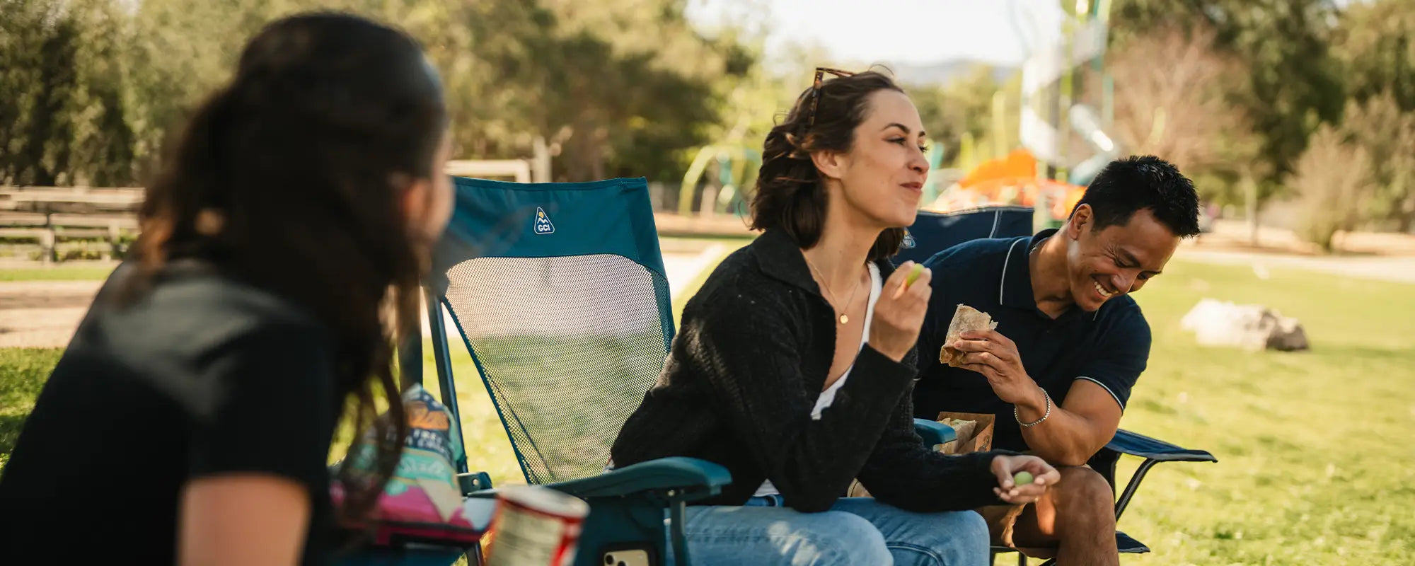 A couple eating snacks at the park as they sit in a BreezeRocker and a Comfort Pro Rocker. 