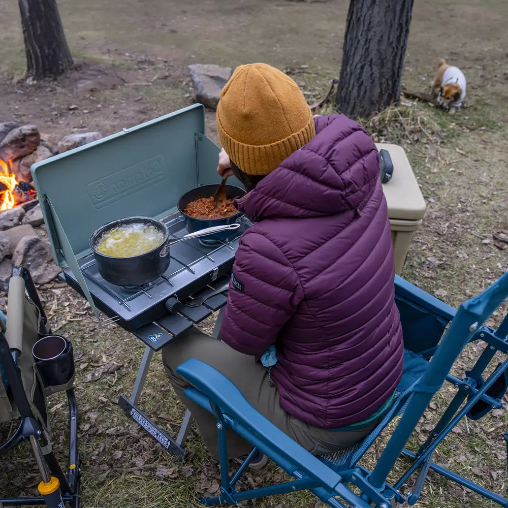 Person seated in a Teal Breeze Rocker cooking at the side table in a campsite setting.