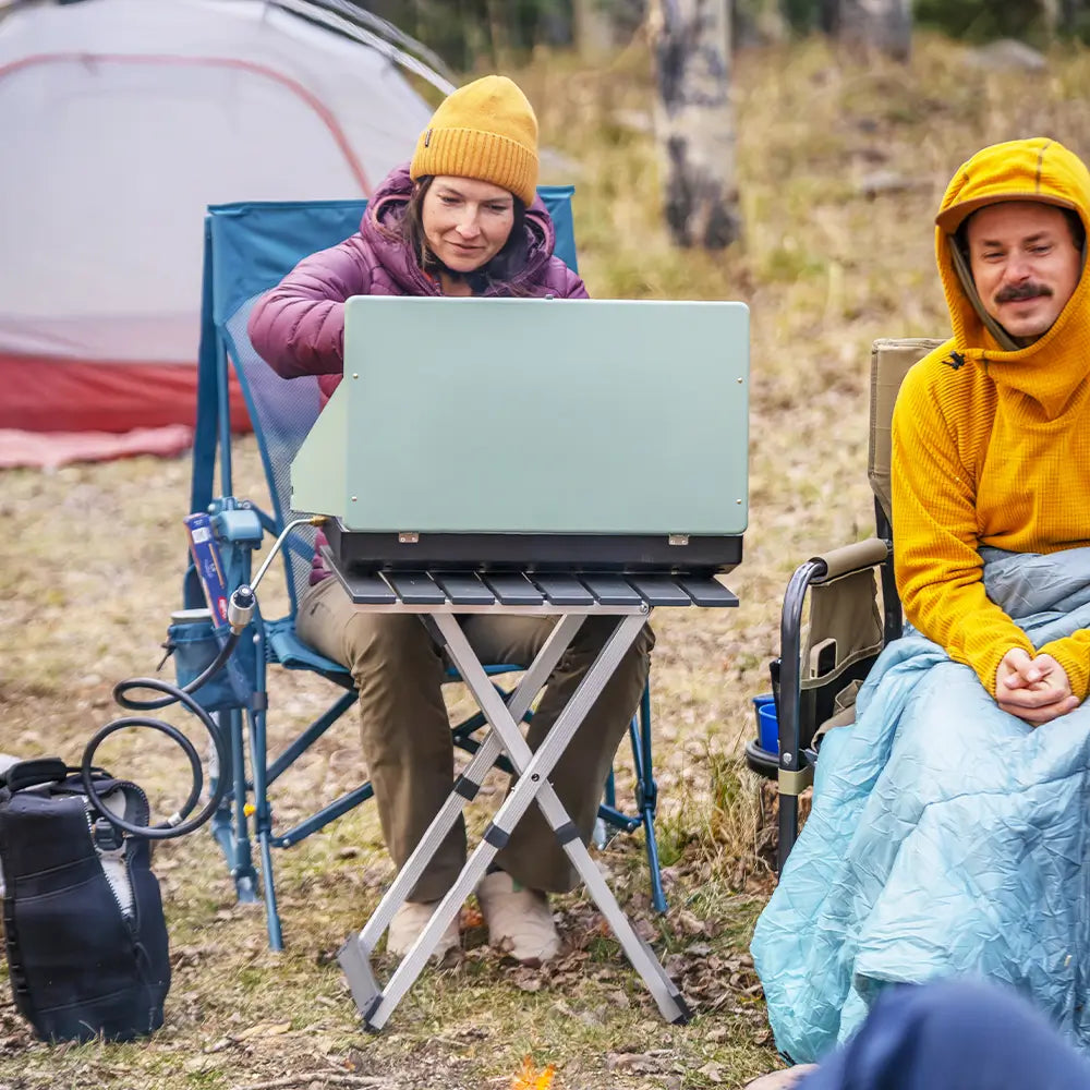 Two people at a campsite using a cooking stove beside a Teal Breeze Rocker.