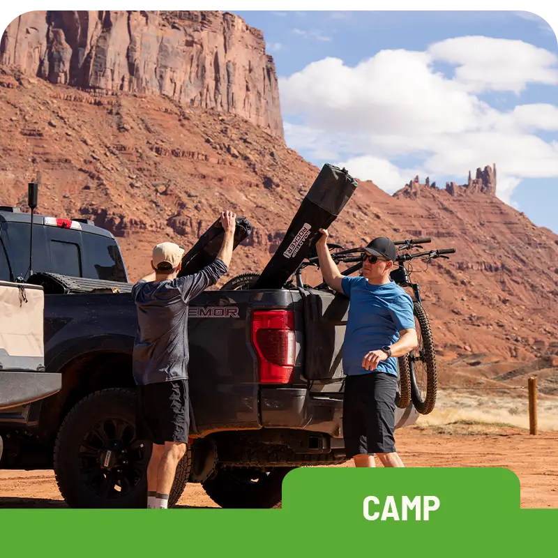 Two men loading outdoor gear into the back of a pickup truck with red rock formations in the background. "Camp" label at the bottom.