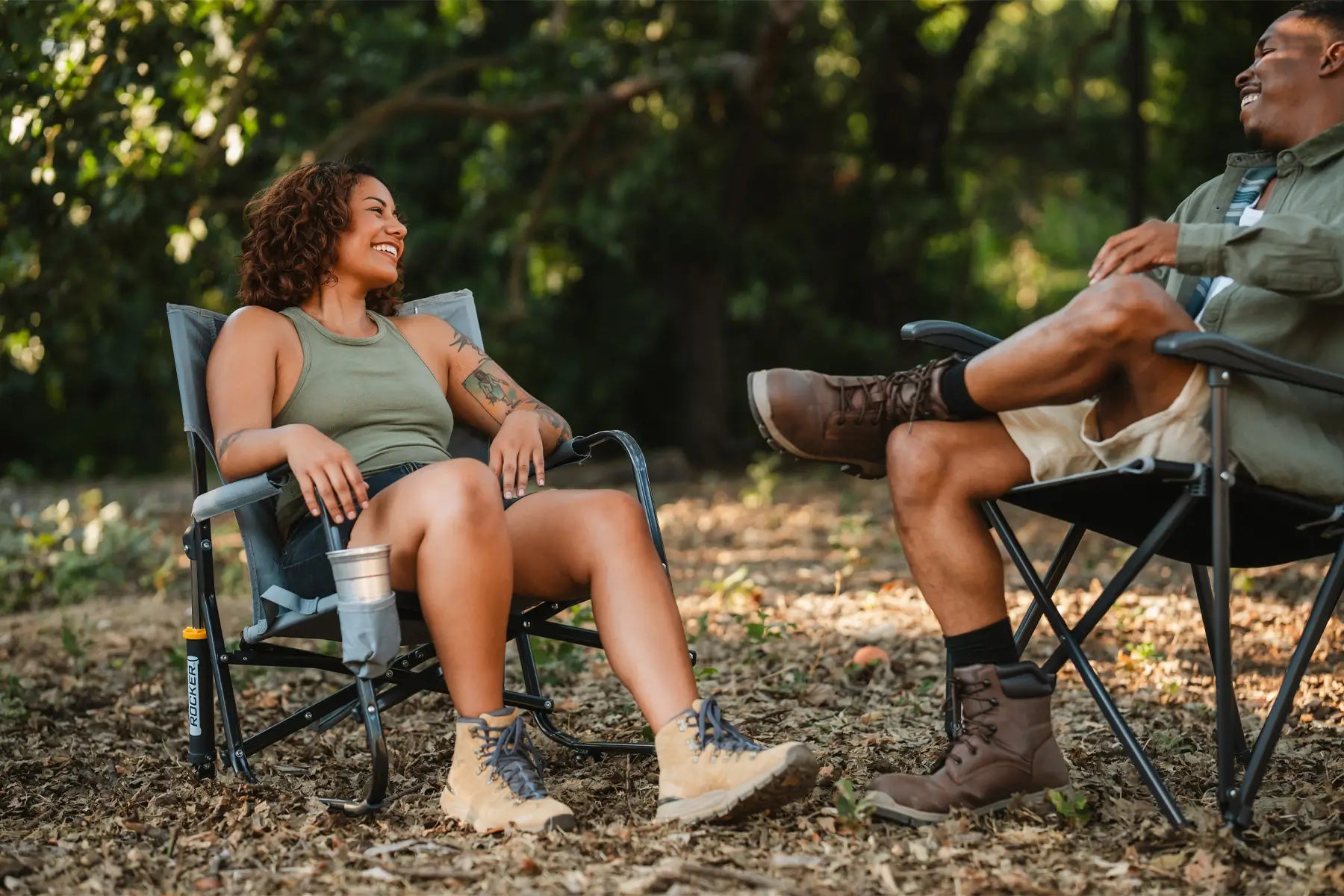 Woman sitting in a Firepit Rocker, laughing with a man in a camping chair, surrounded by trees and fallen leaves.