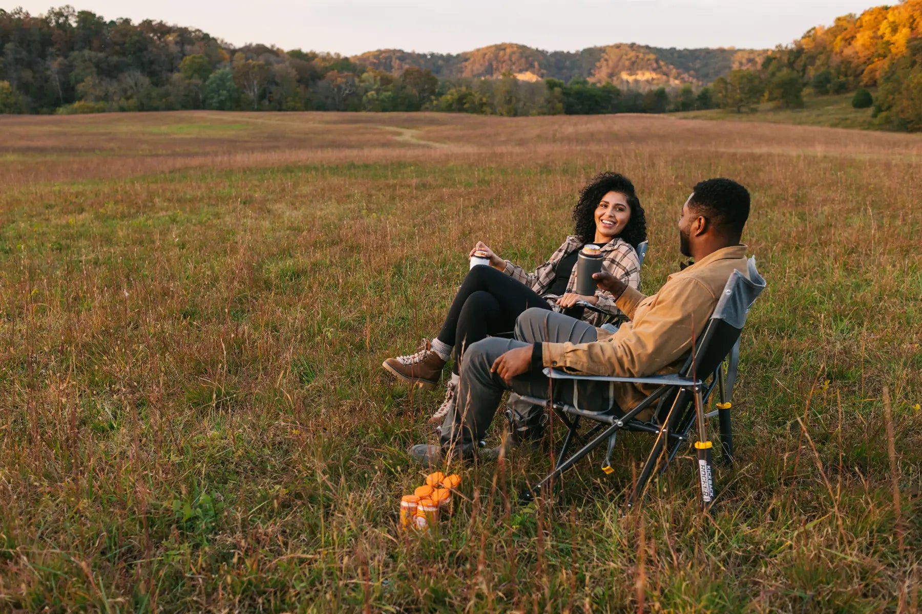 Woman sitting in a kickback rocker chair, laughing with a man in a kickback rocker chair, surrounded by trees and fallen leaves.