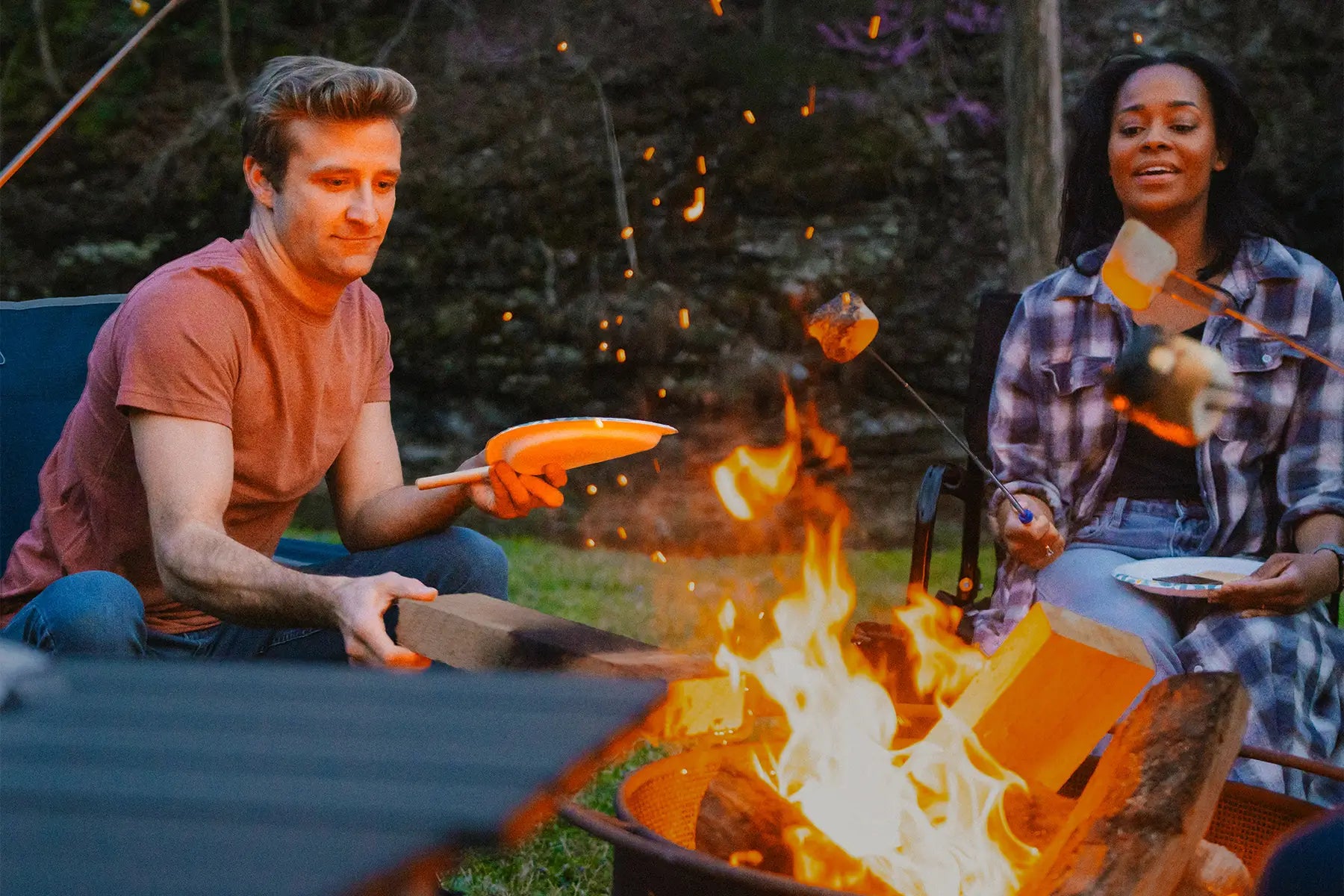 A man and woman roasting marshmallows over a fire while sitting in GCI Outdoor chairs. 