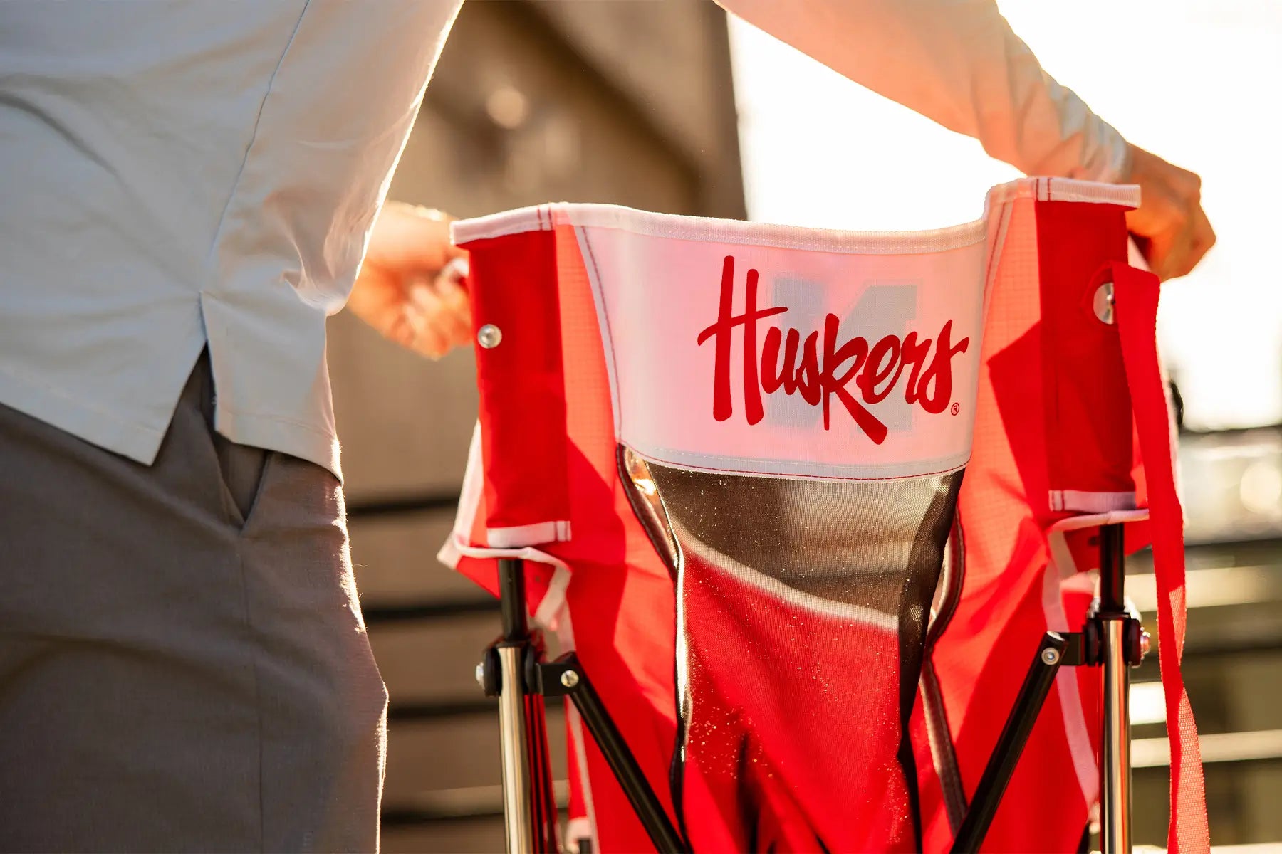 A man unfolding the Nebraska Huskers Comfort Pro Rocker chair showing the back of the chair with a printed "Huskers" on the back. 
