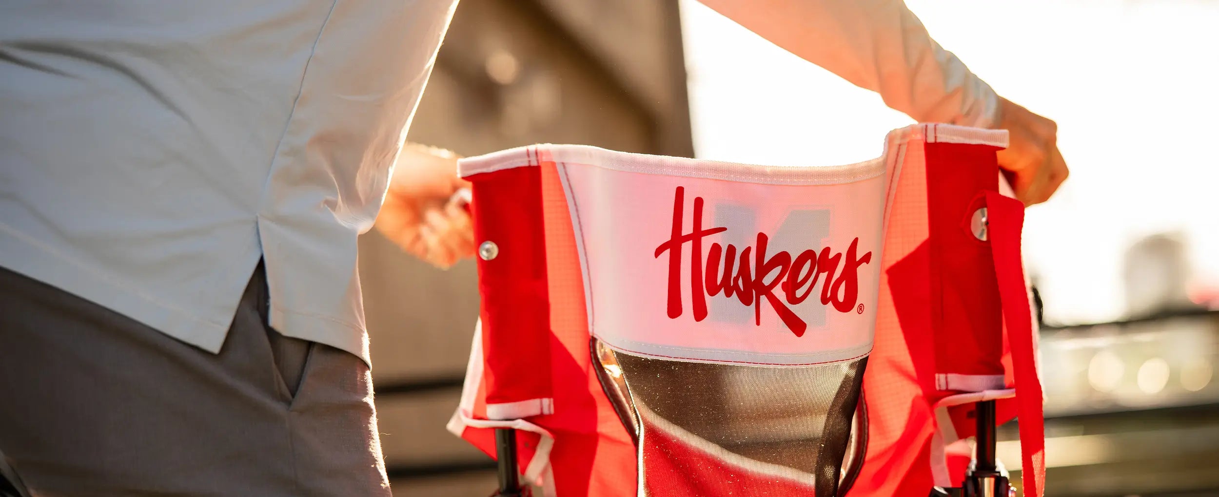 A man unfolding the Nebraska Huskers Comfort Pro Rocker chair showing the back of the chair with a printed "Huskers" on the back. 
