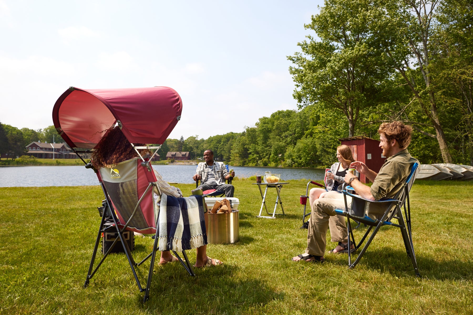 Friends relax by a lake, sitting in Comfort Pro Chairs while chatting and enjoying drinks. A red canopy chair provides shade in the background.