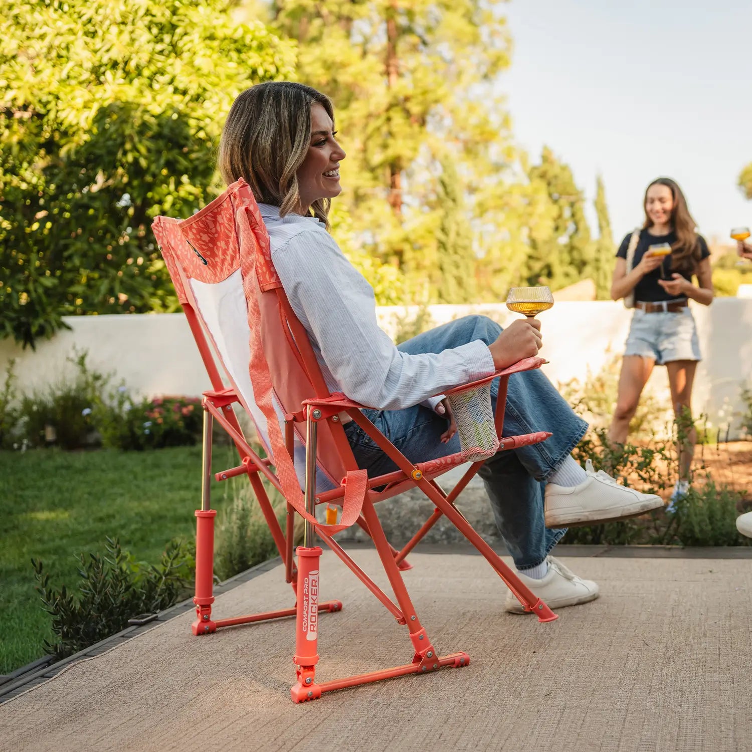 Woman relaxing with a drink in a GCI Outdoor Comfort Pro Rocker in Coral Garden, left side view, at a backyard gathering.