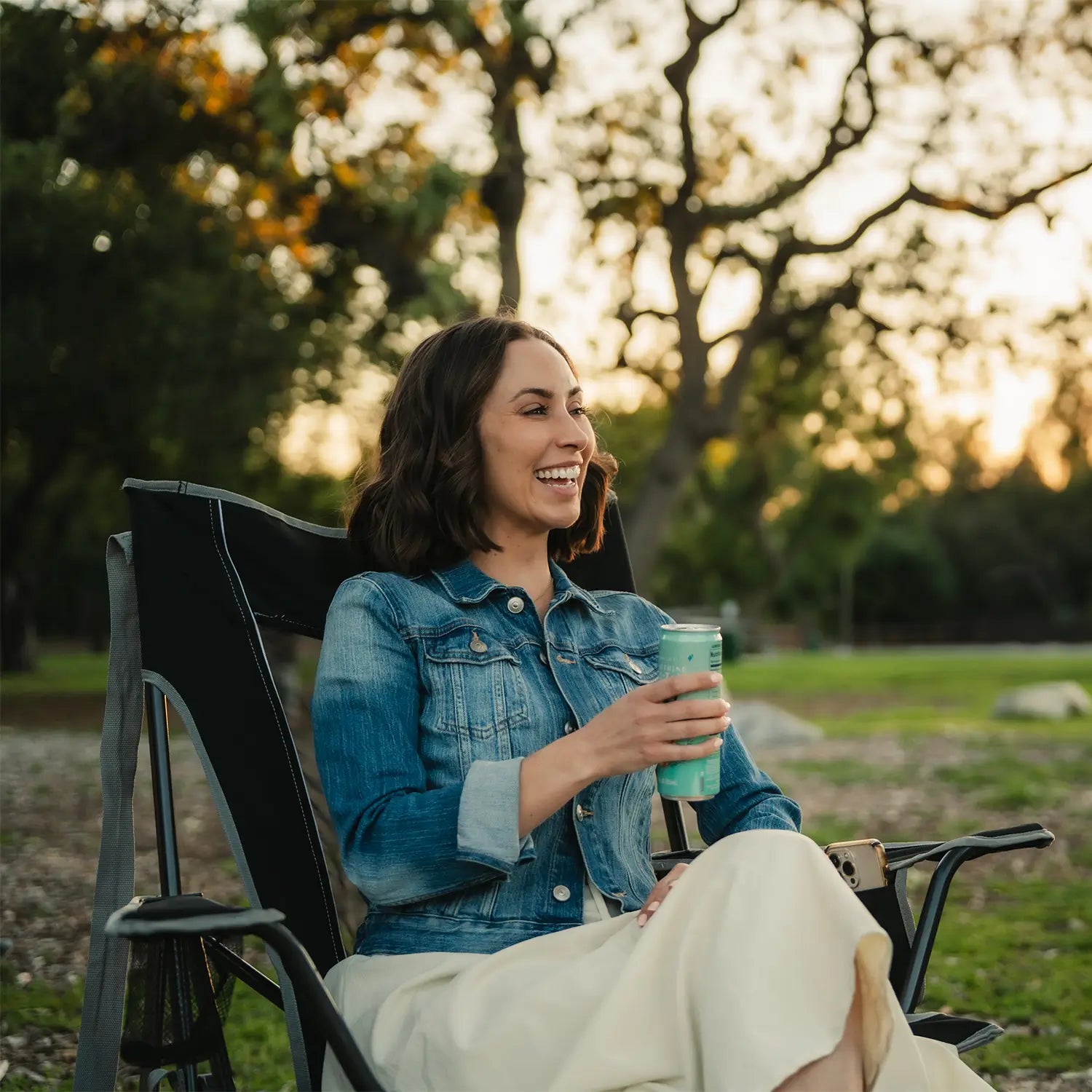 A woman holding a beverage and laughing as she sits in a black Comfort Pro Rocker at a park. 