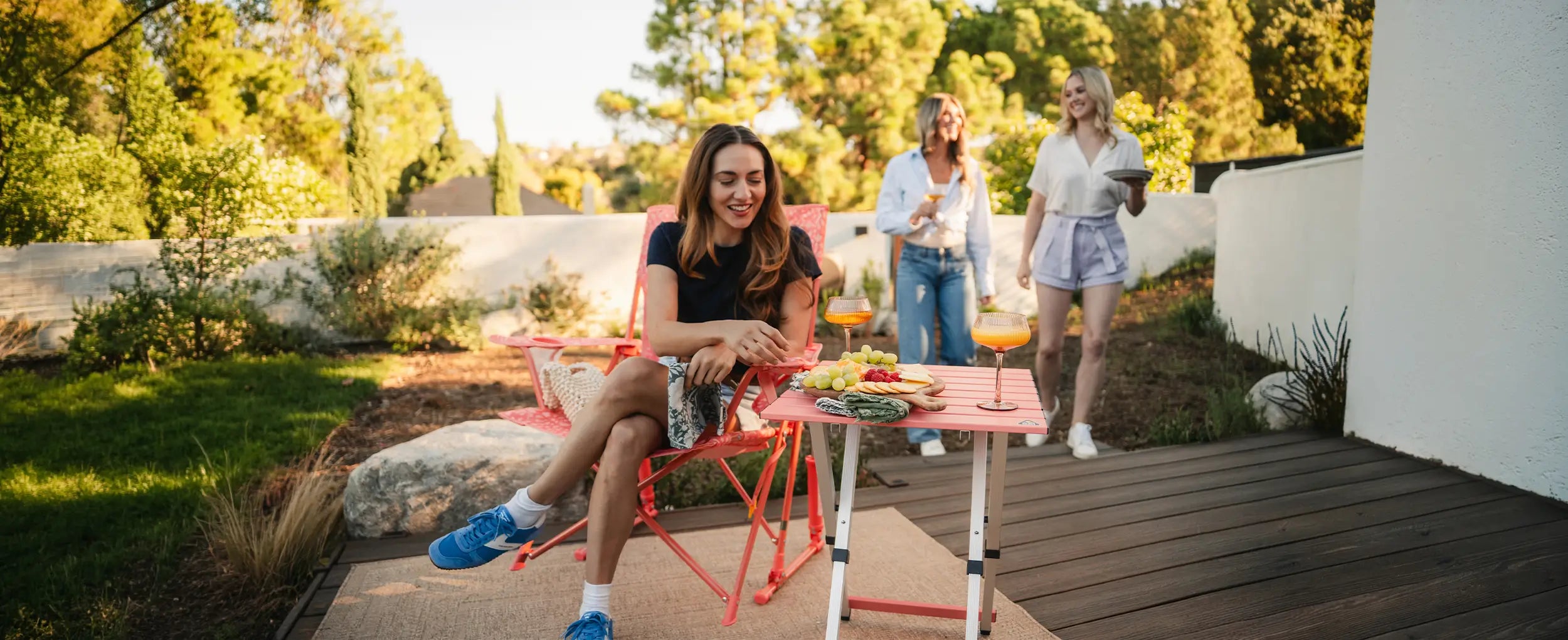 A woman reaching for a snack resting on a coral Compact Table as the woman sits in a coral Comfort Pro Rocker. 