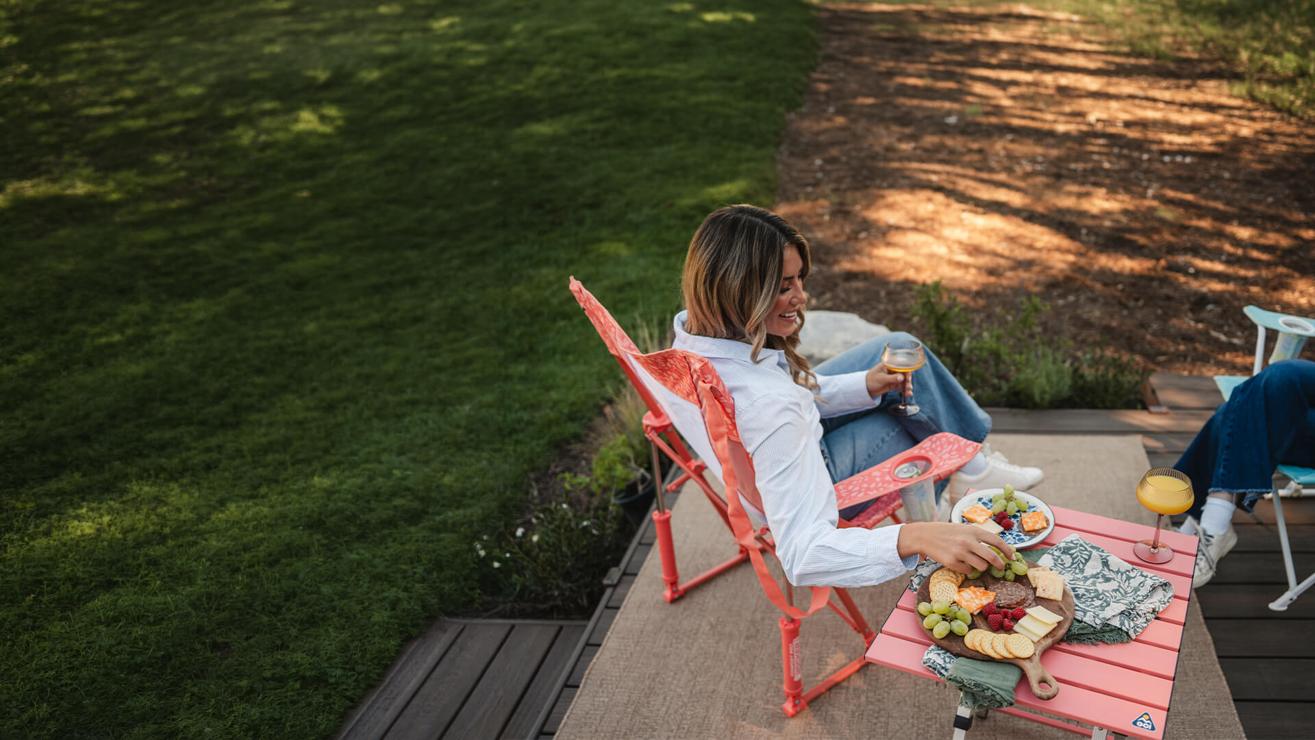 A woman reaching for a snack thats on a coral Compact Camp Table while she sits in a coral Comfort Pro Rocker. 