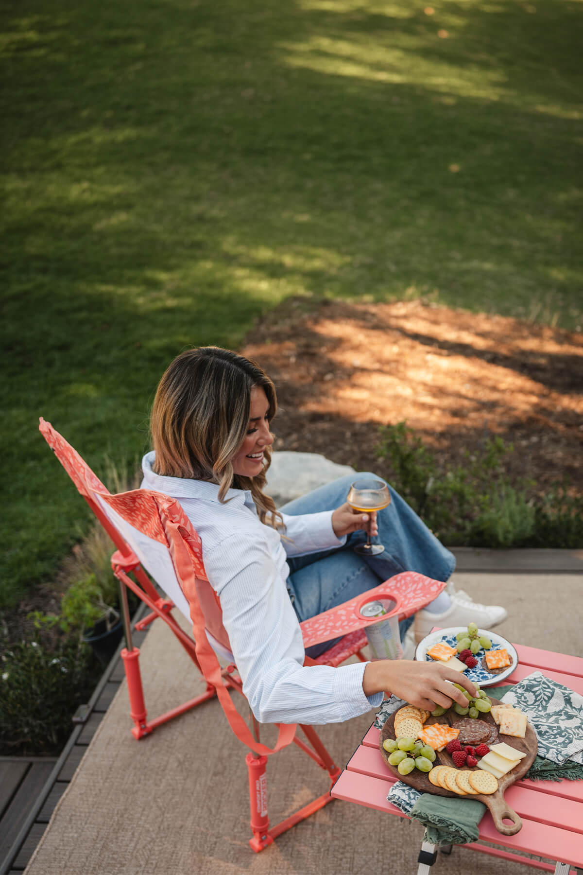 A woman reaching for a snack thats on a coral Compact Camp Table while she sits in a coral Comfort Pro Rocker. 
