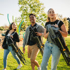 Three people walking outdoors in a park while carrying folded comfort pro rockers on their backs. The setting features green grass and trees under bright daylight.