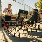 A person in a baseball uniform stands beside a chain-link fence holding a water bottle next to indigo comfort pro rockers, with another person reclining in one of the chairs in an outdoor field setting.