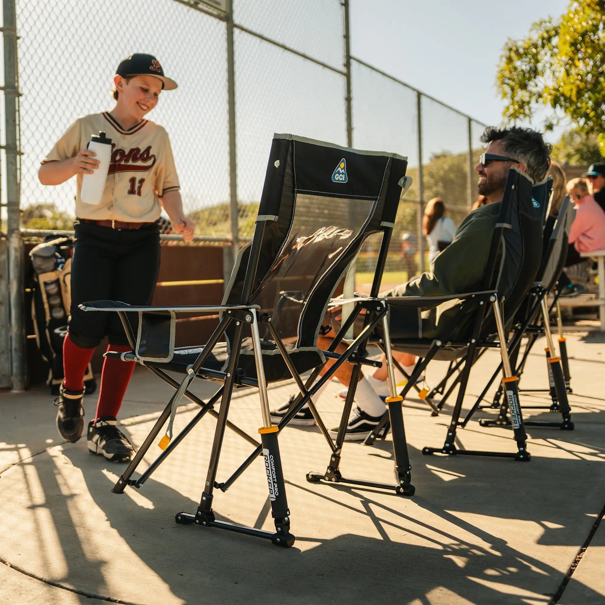 A person in a baseball uniform stands beside a chain-link fence holding a water bottle next to indigo comfort pro rockers, with another person reclining in one of the chairs in an outdoor field setting.