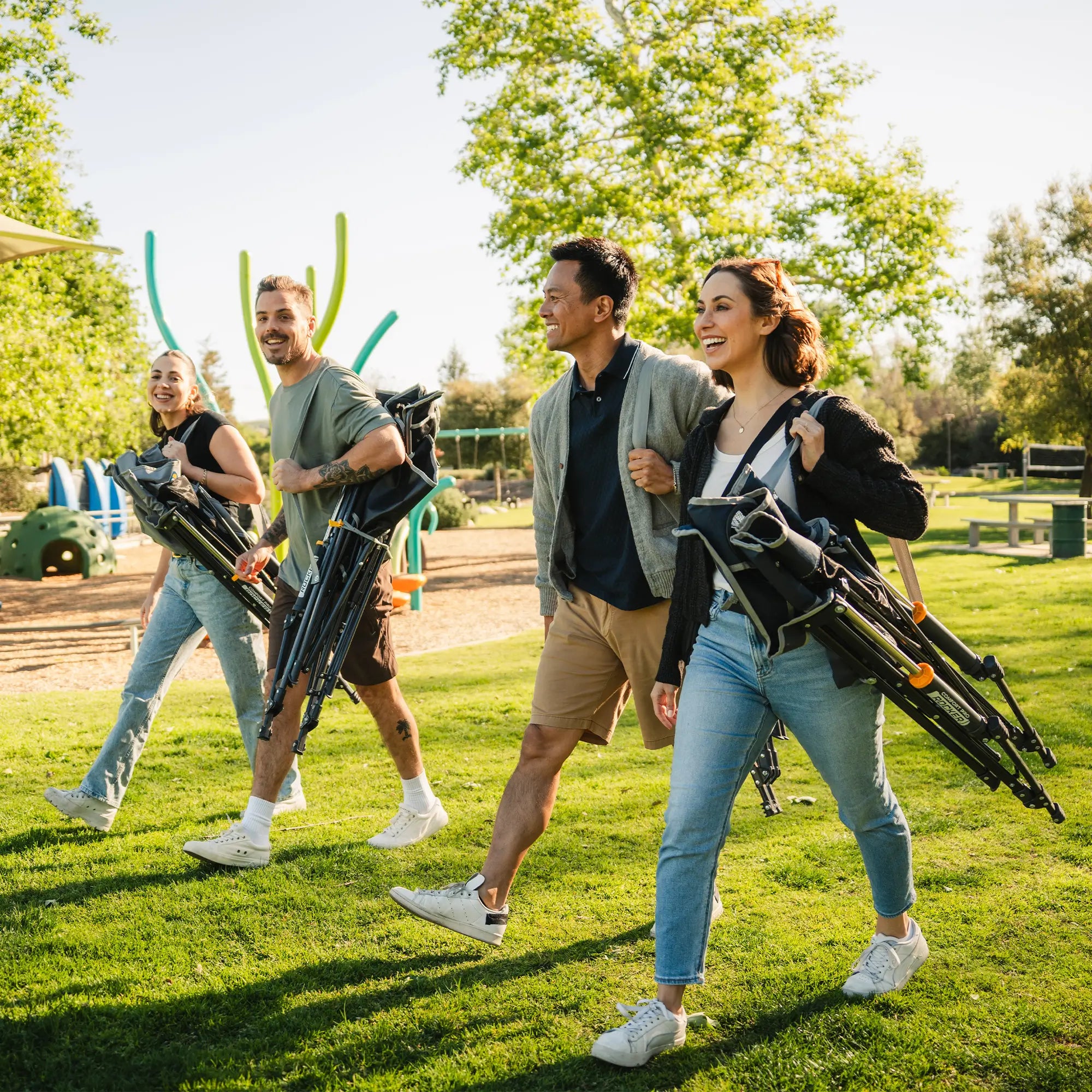 Four people walking outdoors on a sunny park lawn while carrying folded comfort pro rockers. Trees, grass, and playground equipment are visible in the background.