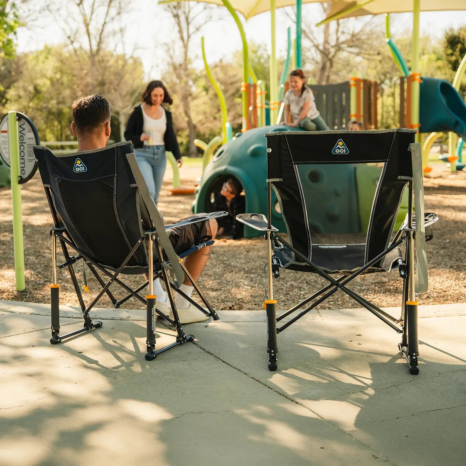 A man sitting in a Comfort Pro Rocker Chair watching his children play on a playground. 