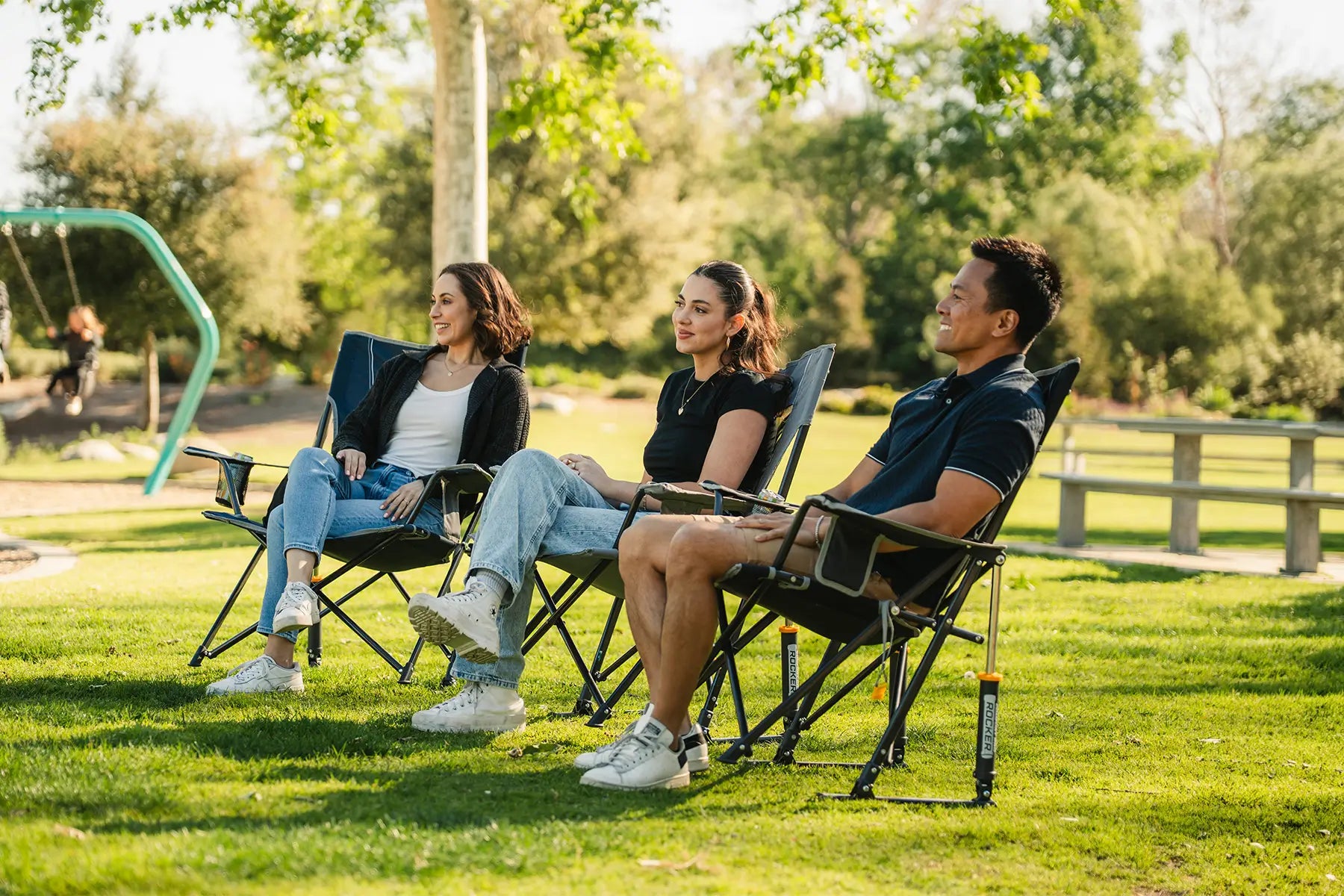 Three people sitting in Comfort Pro Rockers on a grassy park lawn, with trees and a playground visible in the background.